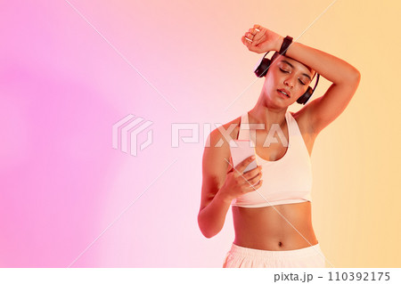 A focused young athlete in workout attire checks her phone for music or fitness app updates while taking A focused young athlete in workout attire checks her phone for music or fitness app updates while taking 110392175