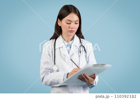 Focused woman doctor writing on clipboard on blue background 110392182