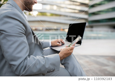 Unrecognizable young businessman using his smartphone while working on blank laptop outdoors 110392183