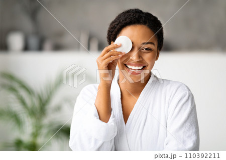 Joyful black woman removing makeup, covering eye with cotton pad 110392211