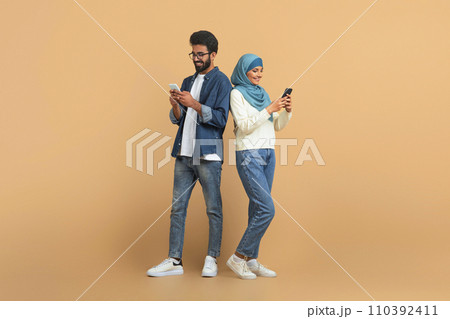 Modern arab muslim couple with their smartphones standing over beige background Modern arab muslim couple with their smartphones standing over beige background 110392411