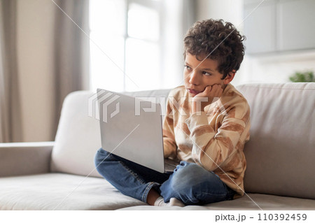 Pensive little black boy sitting with laptop on couch and looking away 110392459