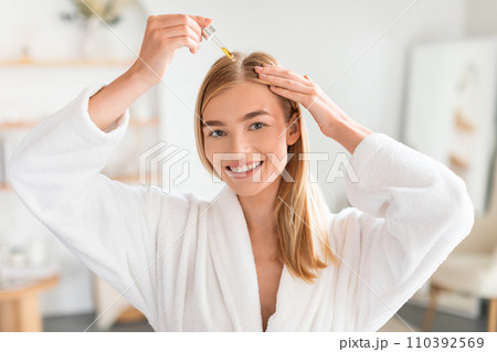 Joyful woman treats hair roots applying nourishing serum in bathroom Joyful woman treats hair roots applying nourishing serum in bathroom 110392569