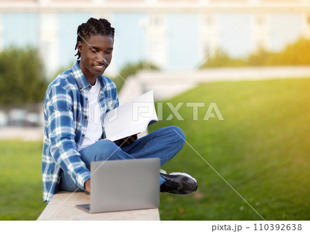 African American student guy websurfing on laptop holding workbooks outside 110392638