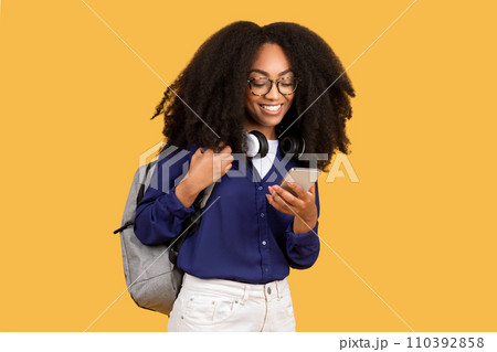 Content black lady student with smartphone and headphones on yellow background 110392858