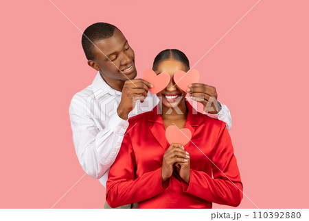 Black man playfully covering woman's eyes with paper hearts, both smiling 110392880