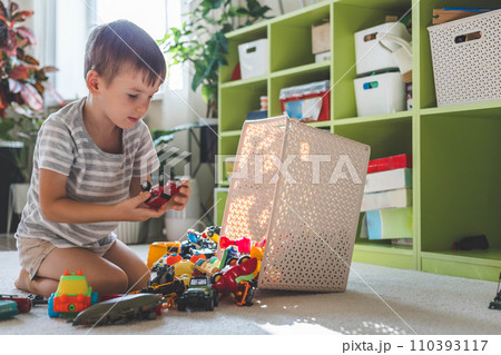 A cheerful European boy plays with cars on the carpet in his room.  110393117