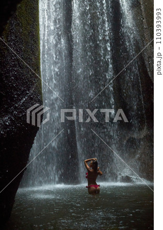 A young, slender woman in a pink swimsuit swims in an incredibly beautiful mystical waterfall in a cliff on the popular island of Bali. 110393993