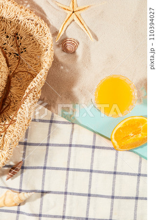 View from above of a glass of fresh orange juice, starfish and shells lying on white sand, a straw hat and a plaid tablecloth. Copy space for ads. View from above of a glass of fresh orange juice, starfish and shells lying on white sand, a straw hat and a plaid tablecloth. Copy space for ads. 110394027