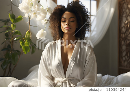 The image conveys peace of mind. A beautiful young woman is meditating on a white bed at home. Dressed in a white bathrobe. luxury clothes. The sun shines down, the fabric flows, its timeless beauty. The image conveys peace of mind. A beautiful young woman is meditating on a white bed at home. Dressed in a white bathrobe. luxury clothes. The sun shines down, the fabric flows, its timeless beauty. 110394244