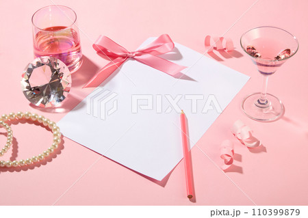 Lovely style scene featured blank paper with a bow and crayon placed on. A cocktail glass and a cup of wine displayed with a diamond and necklace. Copy space 110399879