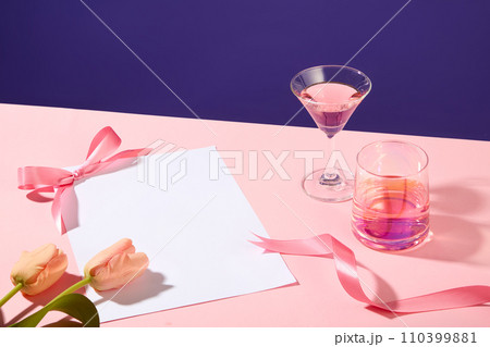 Pink surface featured two flowers, two glasses filled with pink wine and pink ribbons. Lovely or adorable concept. A paper in white color with empty space for text adding 110399881