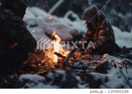 Vibrant campfire crackles amidst a snowy scene, with golden bokeh lights and two blurred figures seated behind, evoking warmth in the cold winter ambiance. 110400286