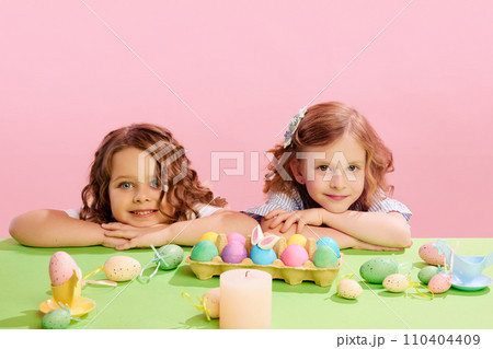 Portrait of beautiful little girls lying on table with decorated, painted eggs against pink background. Easter, childhood 110404409