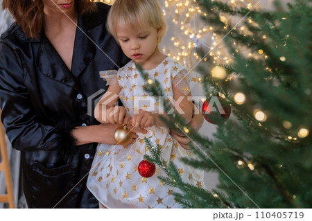 A mother with a 2-year-old daughter decorates the Christmas tree. Mom in a black suit, a girl in a white dress, her daughter hangs a red ball on the Christmas tree. Merry Christmas and New Year A mother with a 2-year-old daughter decorates the Christmas tree. Mom in a black suit, a girl in a white dress, her daughter hangs a red ball on the Christmas tree. Merry Christmas and New Year 110405719