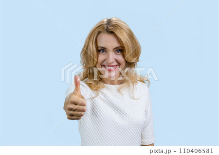 Portrait of a cheerful mature caucasian blond woman gives her thumb up. Isolated on pale blue. 110406581