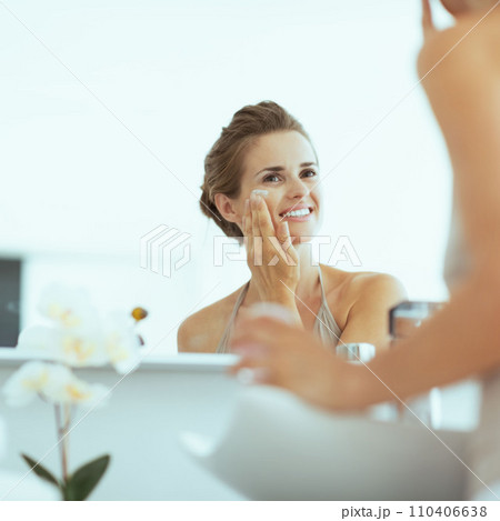 Happy young woman applying cream in bathroom 110406638