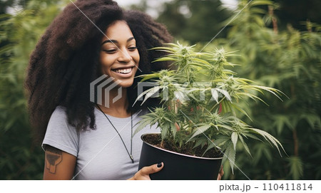 A woman holds a pot of flowering cannabis against a green field. 110411814