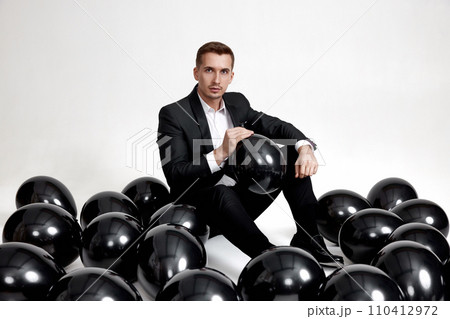elegant caucasian man in suit tuxedo sitting on studio floor with a lot of black air balloons elegant caucasian man in suit tuxedo sitting on studio floor with a lot of black air balloons 110412972