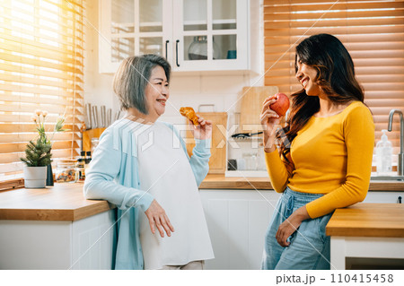 In the kitchen, an attractive young Asian woman and her mother share a moment of togetherness. Their smiles reflect the happiness and family bonding over food and learning. In the kitchen, an attractive young Asian woman and her mother share a moment of togetherness. Their smiles reflect the happiness and family bonding over food and learning. 110415458