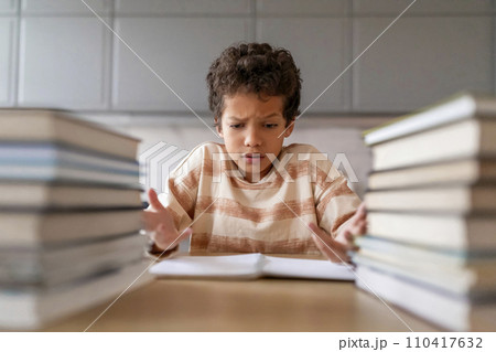 Puzzled black boy making homework surrounded by towering piles of books Puzzled black boy making homework surrounded by towering piles of books 110417632