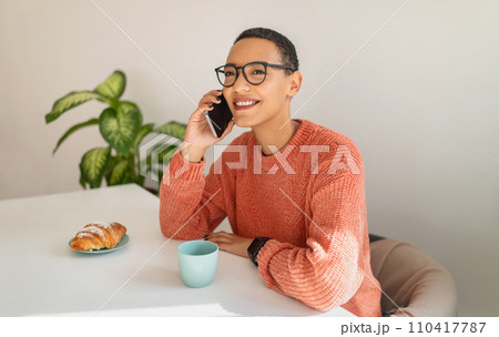 Happy millennial latin lady call by smartphone, drink cup of coffee, eat croissant 110417787