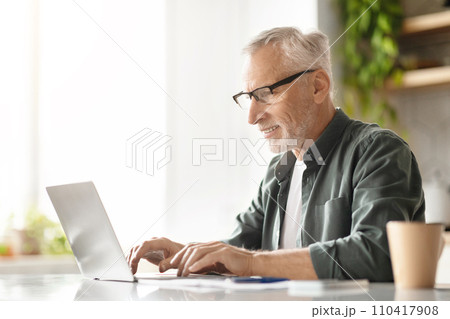 Happy elderly gentleman typing on laptop, enjoying his work in sunny kitchen Happy elderly gentleman typing on laptop, enjoying his work in sunny kitchen 110417908