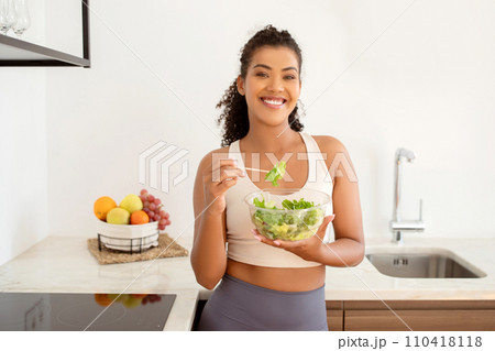 Smiling young lady in sportswear eats vegetable homemade salad indoor 110418118