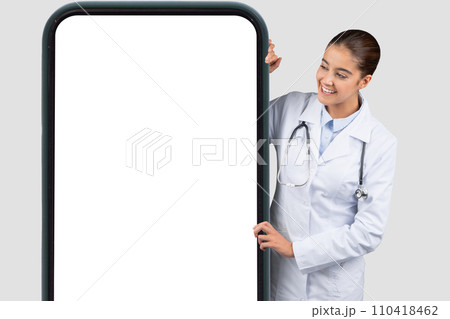 Cheerful Caucasian millennial doctor with a stethoscope appearing from the side of a large smartphone Cheerful Caucasian millennial doctor with a stethoscope appearing from the side of a large smartphone 110418462