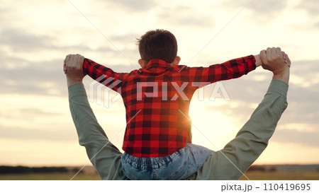 Father holds boy on shoulders spreading arms to sides at sunset on field Father holds boy on shoulders spreading arms to sides at sunset on field 110419695