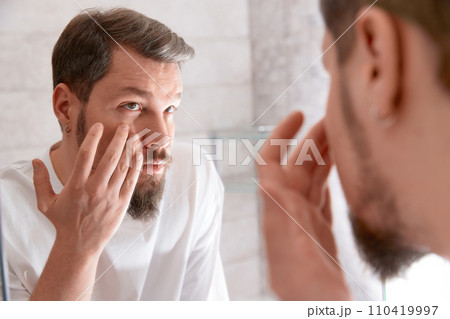 Portrait of man in white tshirt applying cream on his face at bathroom. Self care morning procedure. 110419997