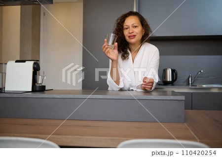 Beautiful young adult woman holding a glass with mineral water, smiling looking at camera, standing at kitchen counter Beautiful young adult woman holding a glass with mineral water, smiling looking at camera, standing at kitchen counter 110420254