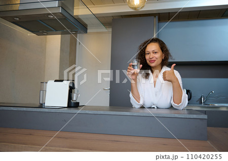 Beautiful curly woman smiles, thumbs up looking at camera, holds a glass of mineral water, standing at kitchen counter 110420255