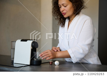 Middle aged Latin woman in white robe, makes fresh espresso using capsule coffee machine, standing in minimalist kitchen 110420263