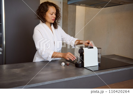 Young woman in white bathrobe, inserting capsule with grinded coffee, preparing espresso in the morning at home kitchen Young woman in white bathrobe, inserting capsule with grinded coffee, preparing espresso in the morning at home kitchen 110420264