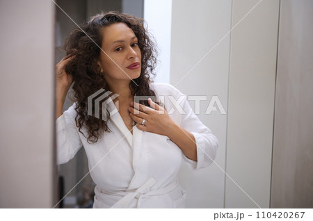 Attractive young adult curly haired brunette woman in white bathrobe, looking at her reflection in the bathroom mirror Attractive young adult curly haired brunette woman in white bathrobe, looking at her reflection in the bathroom mirror 110420267