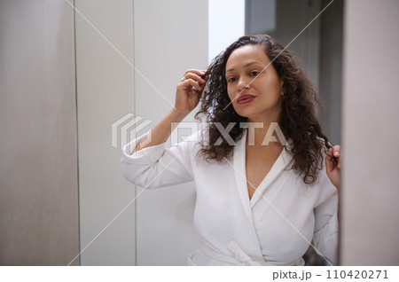 Attractive curly young Caucasian woman in a white robe, holding a strand of hair, looking in the mirror, admiring her appearance, standing in the bathroom at home. Skin, hair and body care concept 110420271