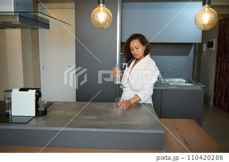 Beautiful African American sleepy woman in white robe holding a glass of refreshing water, standing at kitchen island Beautiful African American sleepy woman in white robe holding a glass of refreshing water, standing at kitchen island 110420286