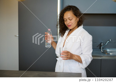 Multi ethnic pretty woman standing at kitchen counter, dressed in white bathrobe, starting her day with glass of water 110420287