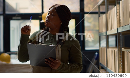 BIPOC warehouse inspector performing inventory audit, keeping quality standards high. Productive expert in distribution center looking at parcels ready to be shipped, crosschecking delivery info 110420699