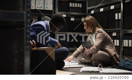 Black policeman and female law officer sitting on office floor with paperwork and laptop as crime investigation is being conducted. Multiracial investigators review and debate case evidence and hints. Black policeman and female law officer sitting on office floor with paperwork and laptop as crime investigation is being conducted. Multiracial investigators review and debate case evidence and hints. 110420700