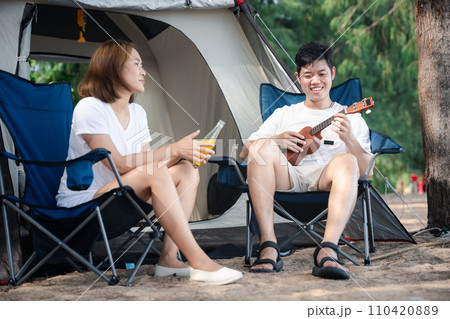 Camping bliss, A cheerful Asian couple, by their tent, serenades each other with ukulele tunes, spreading love and smiles under the setting sun. Togetherness is their melody. Camping outdoor Camping bliss, A cheerful Asian couple, by their tent, serenades each other with ukulele tunes, spreading love and smiles under the setting sun. Togetherness is their melody. Camping outdoor 110420889