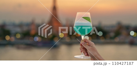 tourist hand shows cocktail glass during enjoy view to Wat Arun Temple in sunset, Traveler visits Temple of Dawn near from rooftop bar. Travel destination in Bangkok, Thailand and Southeast Asia 110427187
