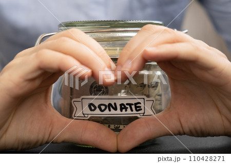 Female showing heart sign with hands Saving Money In Glass Jar filled with Dollars banknotes. DONATE transcription in front of jar. Managing personal finances extra income for future insecurity 110428271