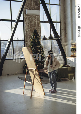 Embraced by the Christmas atmosphere, a young woman in light attire and a VR headset stands before the mirror. 110428621