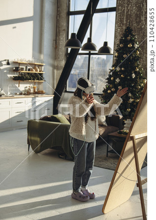 With the Christmas mood around, a young woman in light clothing and a virtual reality headset stands before the mirror. With the Christmas mood around, a young woman in light clothing and a virtual reality headset stands before the mirror. 110428655