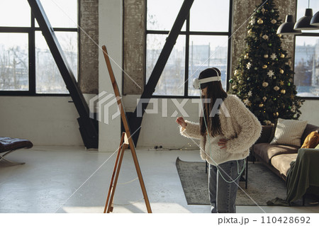 With the holiday ambiance prevailing, a young woman in light attire and a VR headset gazes into the mirror. With the holiday ambiance prevailing, a young woman in light attire and a VR headset gazes into the mirror. 110428692