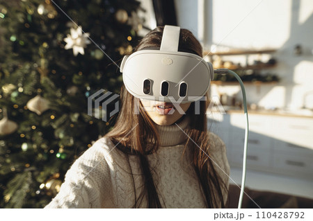 An image of a lovely girl wearing a virtual reality headset against the background of a Christmas tree. 110428792