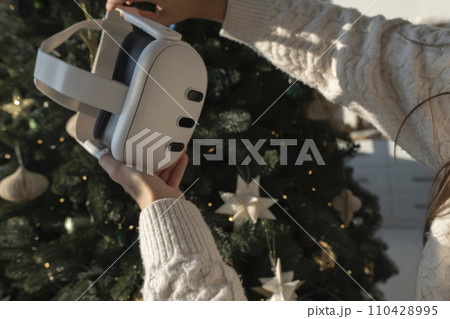 In front of a Christmas tree, a girl is holding a virtual reality headset. 110428995