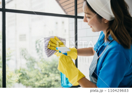 A cheerful woman wearing gloves sprays and cleans office windows. Her dedication to housework emphasizes hygiene and purity ensuring transparent and spotless windows. A cheerful woman wearing gloves sprays and cleans office windows. Her dedication to housework emphasizes hygiene and purity ensuring transparent and spotless windows. 110429683